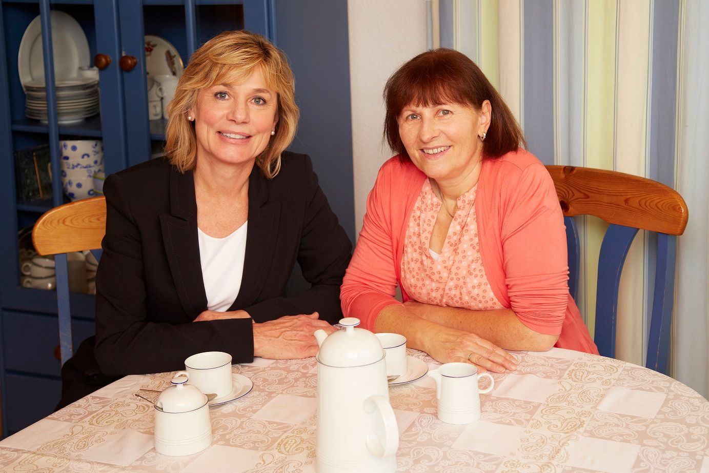 Helga Kröner-Schock sitzt mit ihrer besten Freundin an einem Tisch, beide lächeln in die Kamera. Im Bild eingeblendet ist der Text: „Frau Kröner-Schock trinkt Kaffee mit ihrer besten Freundin.“