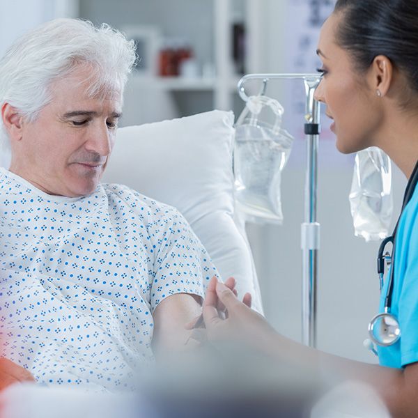 Zu sehen ist links ein im Krankenhausbett aufrecht sitzender Mann im Krankenhauskittel. Rechts daneben befindet sich eine junge Ärztin in sitzender Position, in blauer Krankenhauskleidung und Stethoskop. Im Hintergrund ist ein Ständer mit Tropf zu sehen.