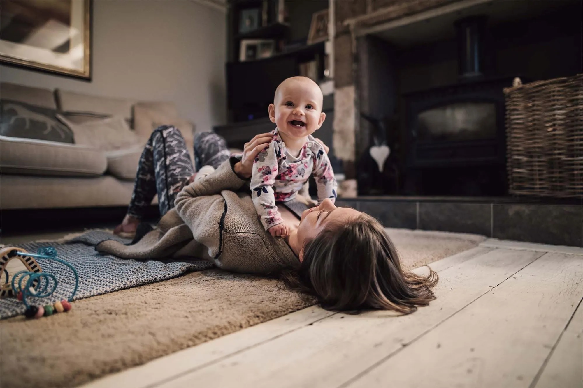 Eine Mutter liegt mit ihrem Baby auf dem Teppich des Wohnzimmers. Das Baby krabbelt auf ihrem Bauch und lacht.