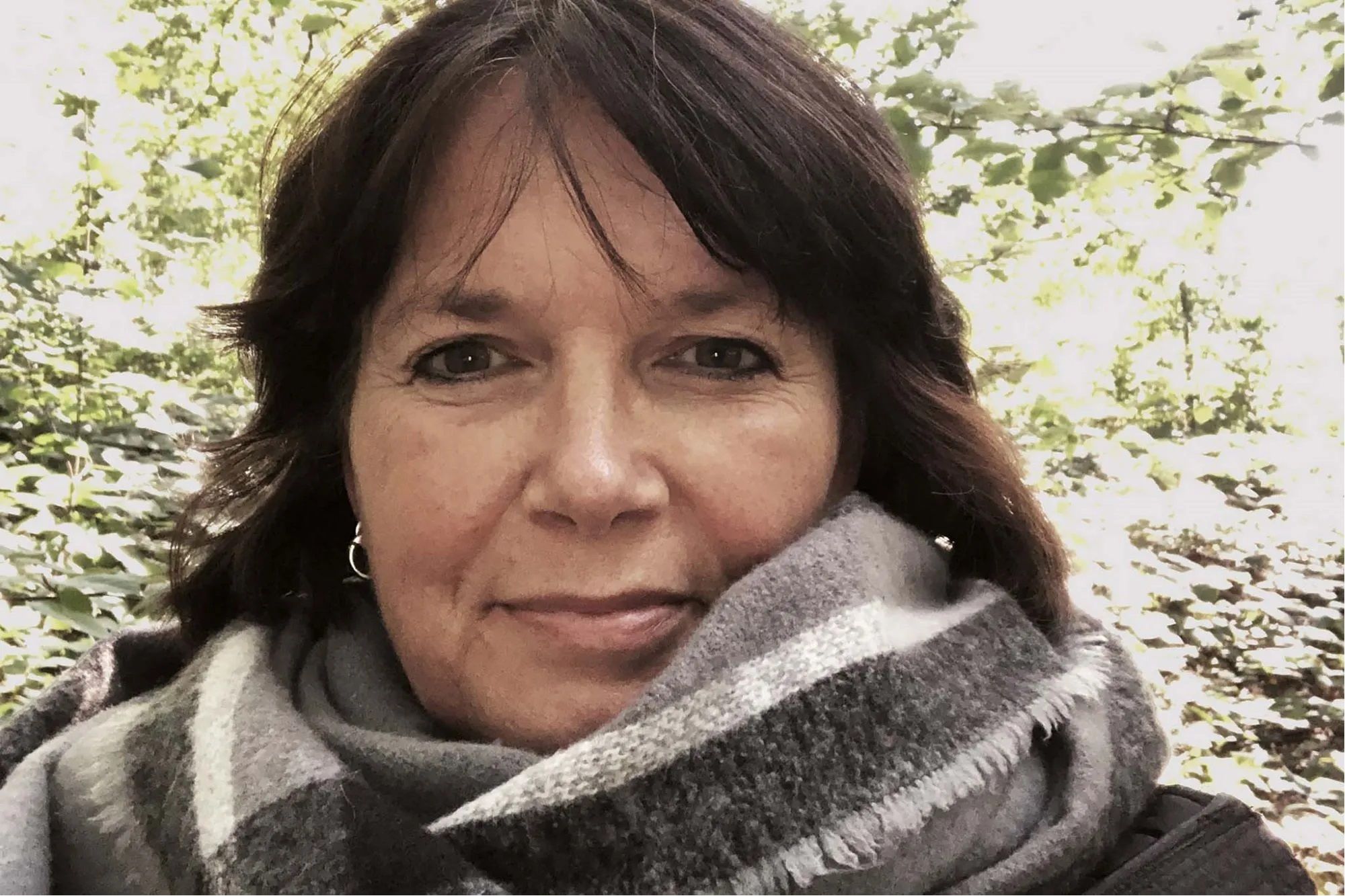 Martina steht im Wald und blickt ruhig in die Kamera. Sie trägt einen dicken Schal und eine Jacke. Um sie herum sind Bäume und herbstliches Laub zu sehen.
