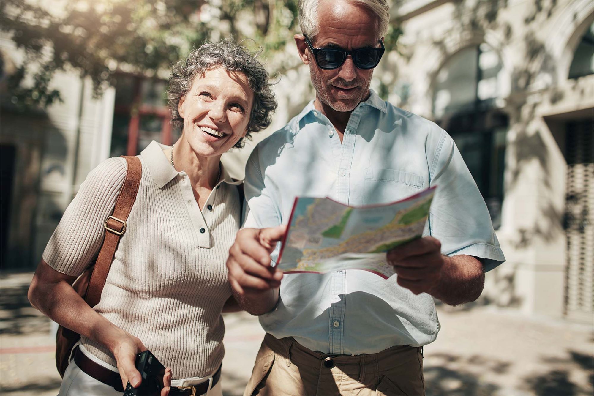 Ein Paar im mittleren Alter flaniert bei Sonnenschein durch eine Stadt. Sie hat kurze graue Locken und hält einen Fotoapparat in der Hand. Sie lacht. Er ist braungebrannt, trägt eine Sonnenbrille und orientiert sich mithilfe eines Stadtplans.