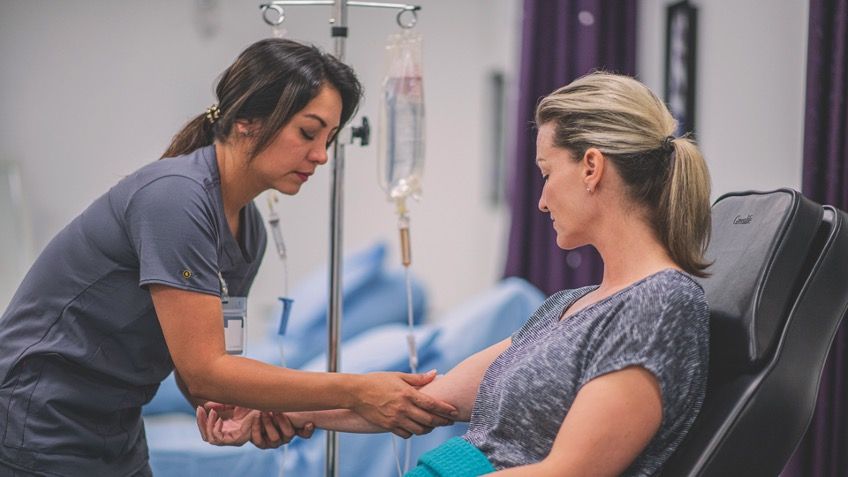 Pflegekraft legt einer auf einem Behandlungsstuhl sitzenden Patientin einen intravenösen Zugang mit Infusion an.