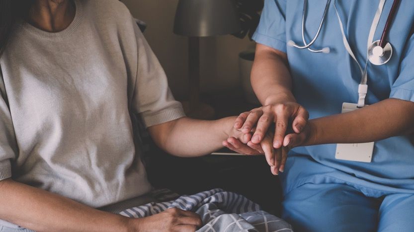 Pflegekraft legt ihre Hand auf die einer sitzenden Patientin.