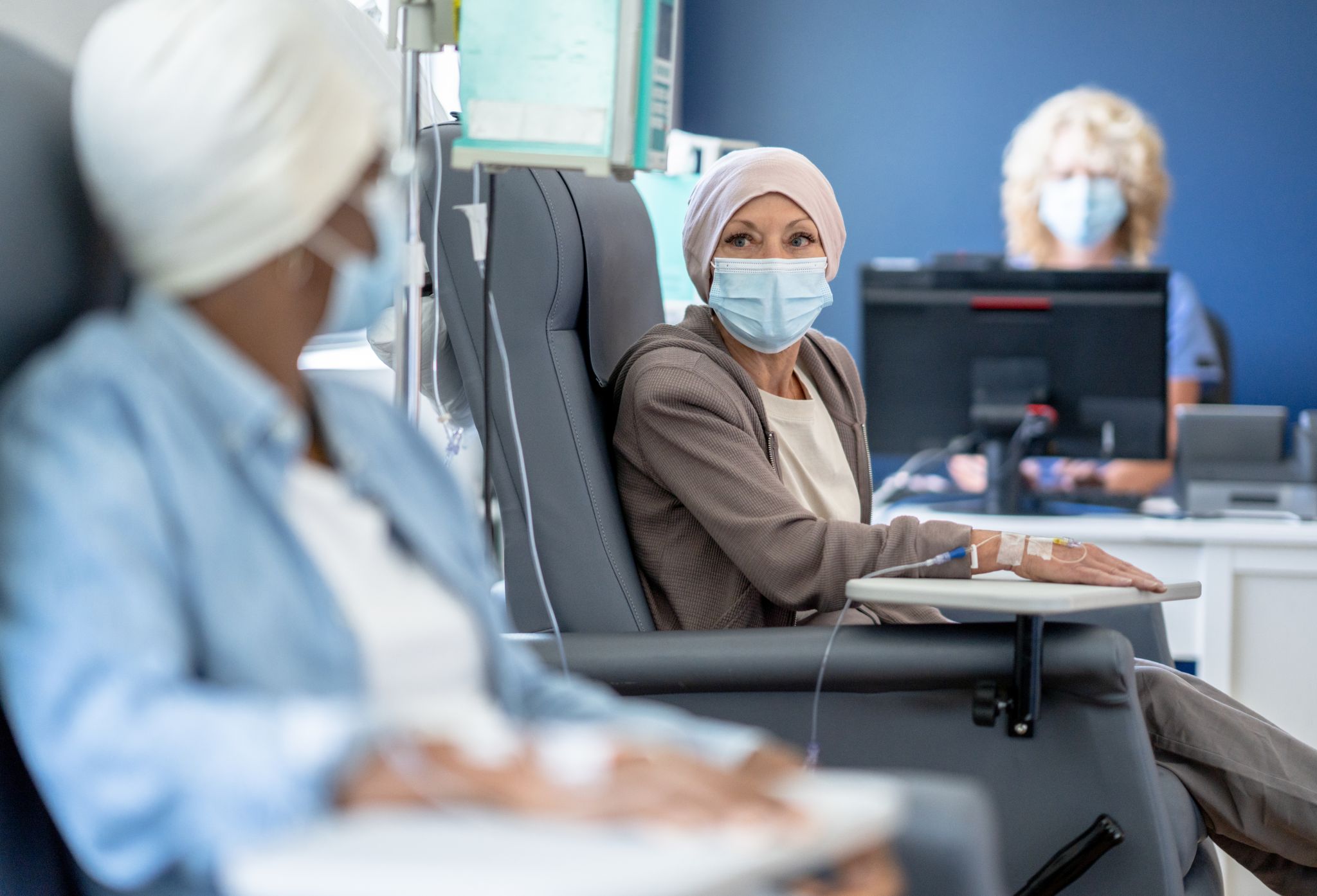 Zwei Patientinnen mit Mund-Nasen-Schutz und Kopftüchern bei der Chemotherapie, im Hintergrund eine medizinische Fachkraft mit Mund-Nasen-Schutz. 