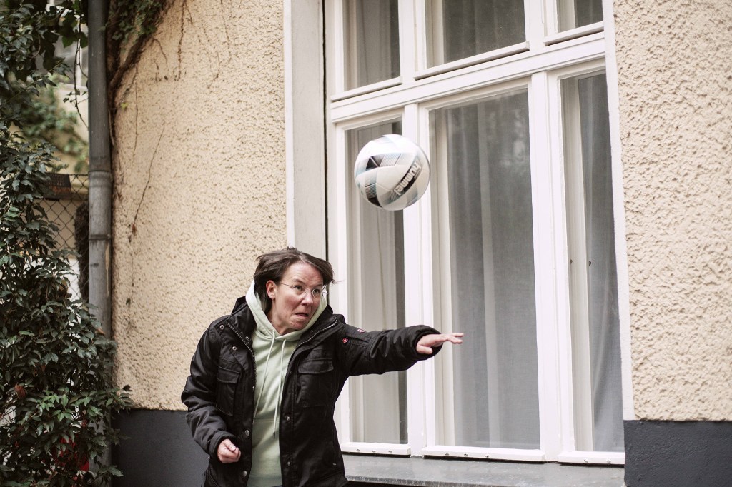 Patrycja steht vor einer Hauswand und spielt konzentriert mit einem Fußball. Sie streckt die Hand aus und trägt eine schwarze Jacke über einen Kapuzenpullover.