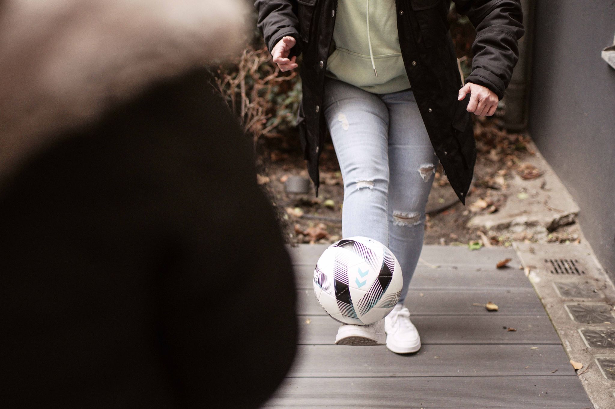 Patrycja spielt draußen auf einem Holzboden mit einem Fußball. Mit Ihrem Bein hält sie den Ball in der Luft. Sie trägt eine graue Jeans, weiße Sneaker, einen grünen Pullover und eine schwarze Jacke.