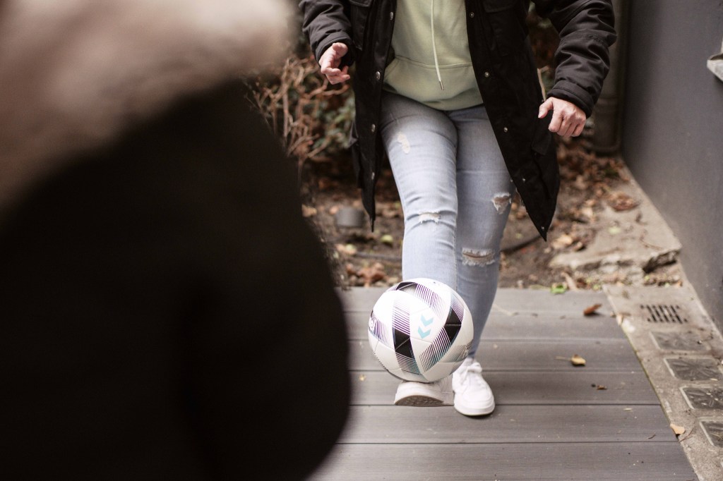 Patrycja spielt draußen auf einem Holzboden mit einem Fußball. Mit Ihrem Bein hält sie den Ball in der Luft. Sie trägt eine graue Jeans, weiße Sneaker, einen grünen Pullover und eine schwarze Jacke.