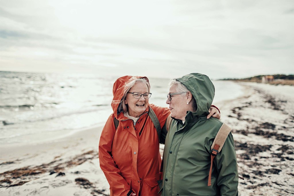 Zwei Frauen im Seniorenalter machen einen Spaziergang am Meer. Der Himmel ist wolkig, das Wetter kühl. Der Strand um sie herum ist menschenleer. Die beiden tragen Regenjacken, eine orange-rot, eine dunkelgrün, und Rucksäcke. Die Frau links in der rötlichen Jacke hat ihren Arm um ihre Freundin gelegt.