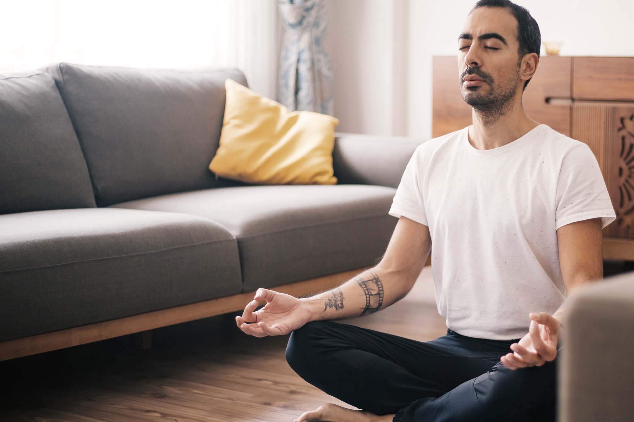 Ein junger Mann meditiert in seinem Wohnzimmer: Er sitzt in bequemer Kleidung mit überkreuzten Beinen im Lotussitz auf einer Yogamatte.