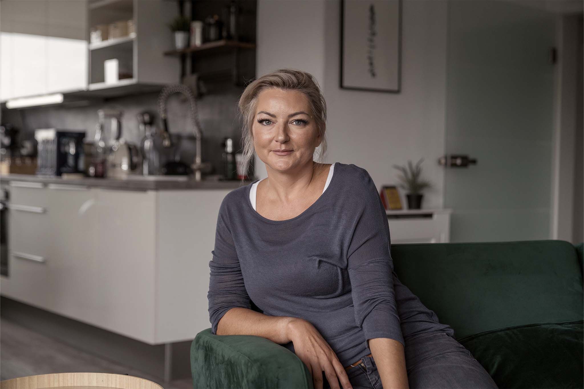Moni sitzt auf eine Armlehne gesetzt auf einem dunkelgrünen Sofa. Sie trägt ein dunkelgraues Oberteil, dessen Ärmel sie bis zu den Ellenbogen hochgekrempelt hat. Ihre blonden Haare hat sie zu einem Zopf gebunden. Im Hintergrund ist die links die Ecke einer Küchenzeile zu sehen.