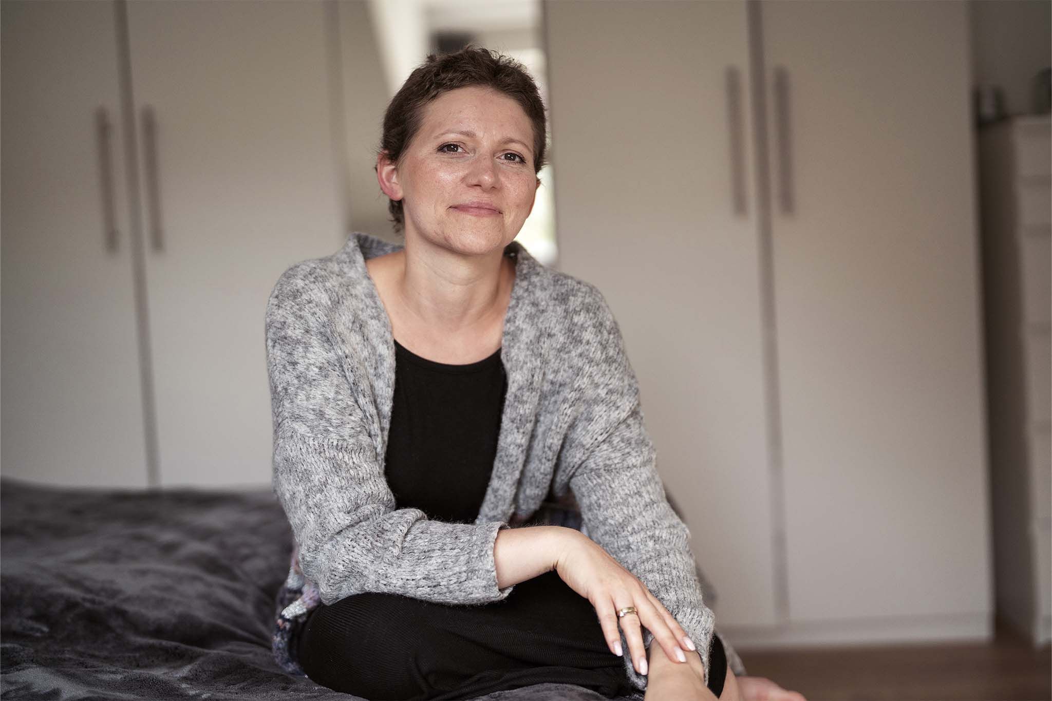 Silja sitzt vor ihrem Kleiderschrank und blick lächelnd in die Kamera. Sie trägt ein schwarzes Oberteil und ein grau-gemusterte Strickjacke. Sie hat kurze, braune Haare und dunkle Augen.