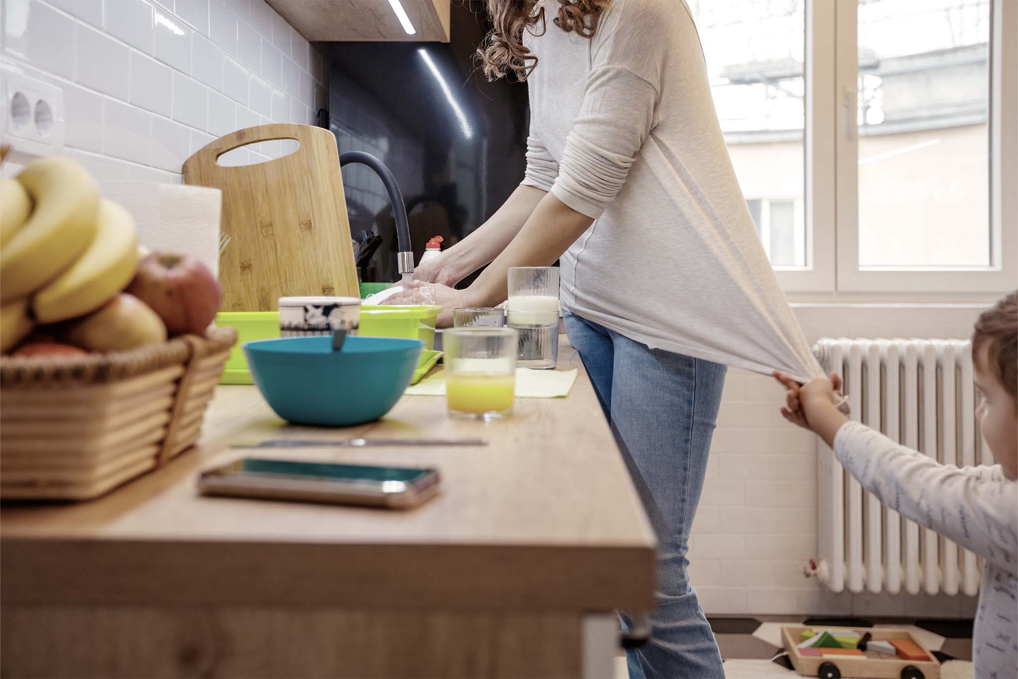 Eine Frau steht in der Küche und macht den Abwasch. Währenddessen zieht ein kleines Kind an ihrem Oberteil und möchte ihre Aufmerksamkeit erregen.