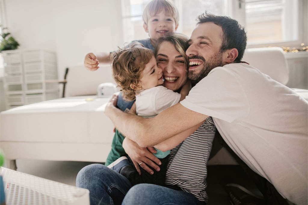 Eine Familie sitzt eng aneinander geschmiegt im Wohnzimmer auf dem Boden. Alle vier lachen. Die Mutter hält ein kleines Mädchen im Arm, hinter ihr breitet ein kleiner Junge die Arme aus. Der Vater umarmt alle drei.