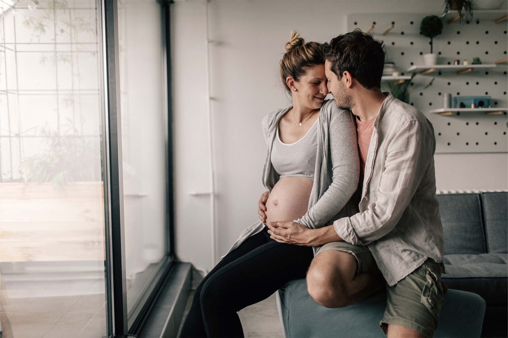 Ein Paar sitzt zuhause am Fenster auf dem Sofa, die Gesichter einander zugeneigt. Die Frau ist schwanger. Er hält sie von hinten umschlungen und streicht über ihren nackten Babybauch.