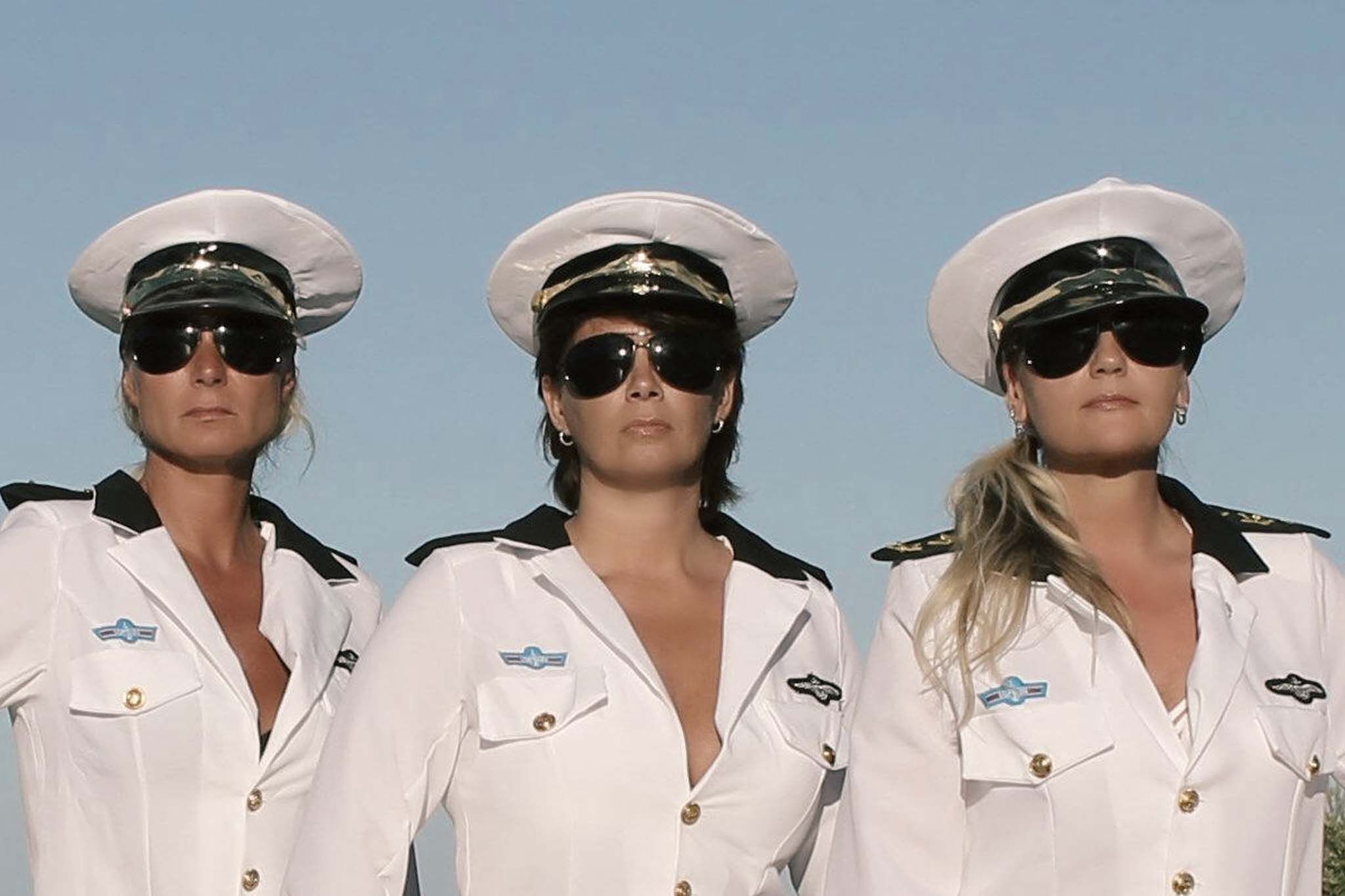 Martina steht mit zwei weiteren Frauen nebeneinander und trägt gemeinsam mit ihnen weiße Kostüme im Stil von Uniformen mit Mützen und Sonnenbrillen. Alle drei blicken ernst in die Kamera, vor einem klaren blauen Himmel.