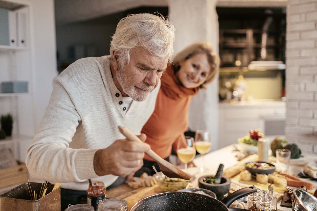 Ein älteres Paar kocht gemeinsam. Vor ihnen liegen verschiedene Zutaten und Kochgeräte. Der Mann beugt sich zum Probieren über eine Pfanne.