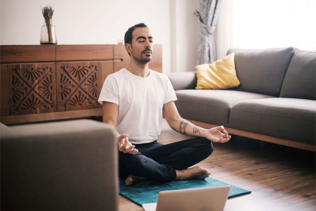 Ein junger Mann meditiert in seinem Wohnzimmer: Er sitzt in bequemer Kleidung mit überkreuzten Beinen im Lotussitz auf einer Yogamatte.