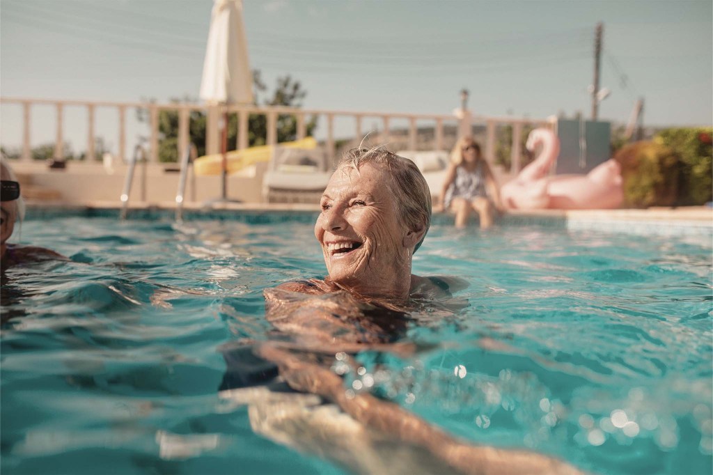 Eine ältere Krebsüberlebende schwimmt in einem Pool unter freiem Himmel. Sie trägt einen blauen Badeanzug und lacht.