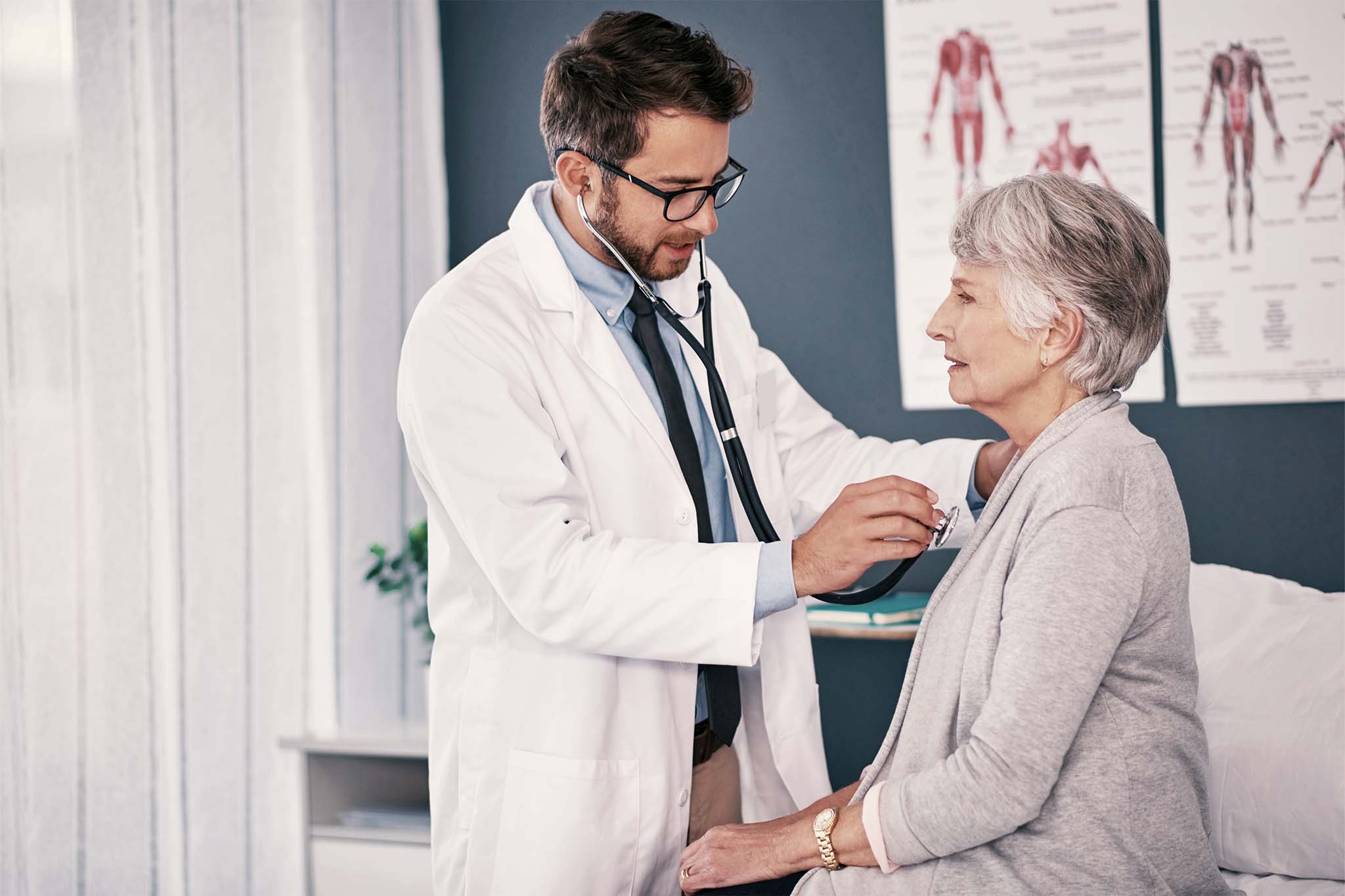 Eine ältere Patientin in einer grauen Jacke sitzt in einer Arztpraxis. Vor ihr steht ihr Arzt, der sie untersucht.