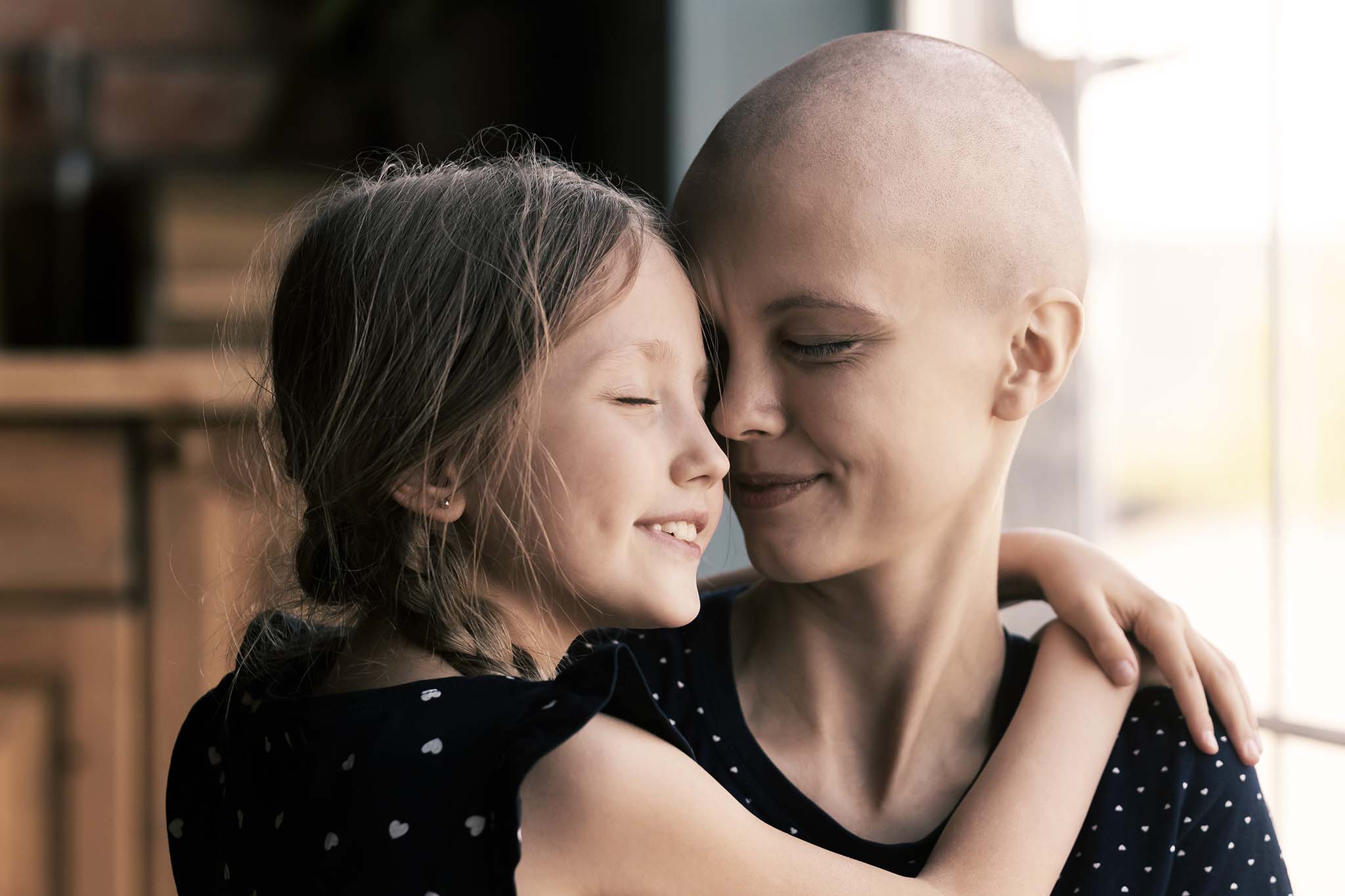 Eine junge Frau nach einer Chemotherapie sitzt mit ihrer Tochter am Fenster. Das kleine Mädchen hat ihre Arme um die Mutter gelegt.
