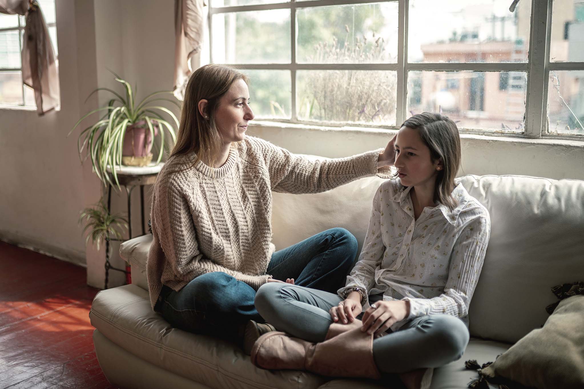Eine junge Frau und ein Mädchen im Teenageralter sitzen auf einem Sofa. Die junge Frau streicht dem Mädchen über den Kopf.