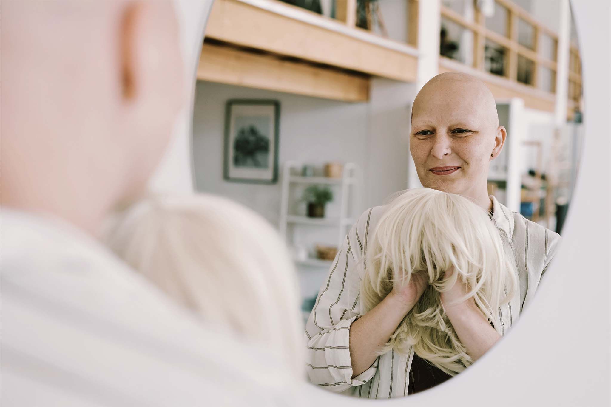 Eine Frau mit Glatze steht vor einem Spiegel. Vor sich hält sie eine blonde Perücke über beide Hände gespannt.