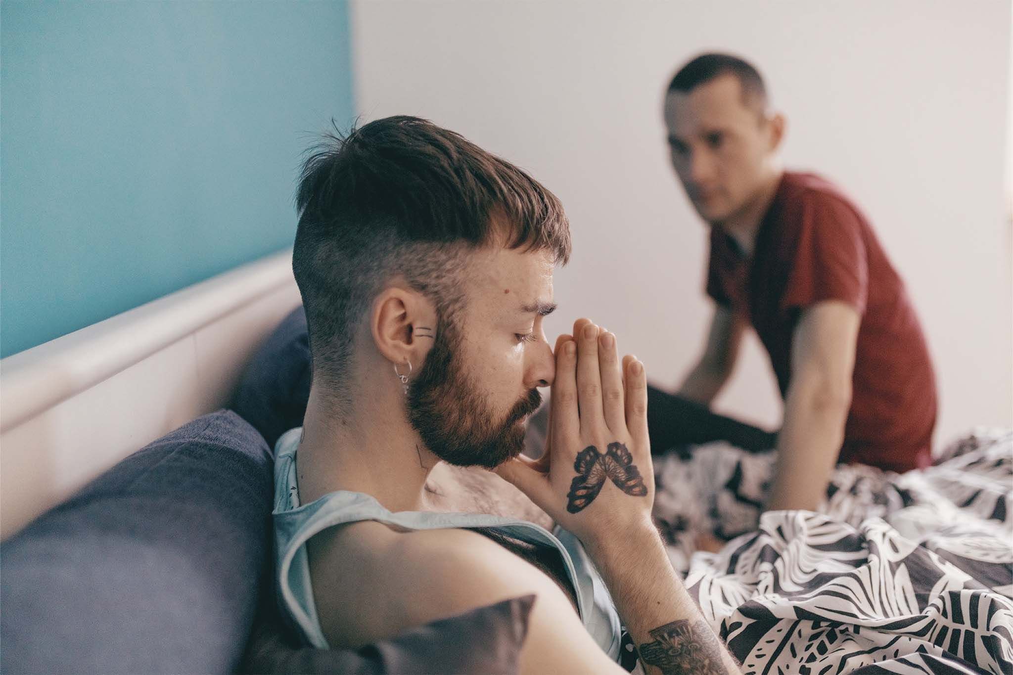 Ein Mann liegt mit vor dem Gesicht zusammengelegten Händen im Bett und hat die Augen geschlossen. Sein Partner sitzt auf der anderen Seite des Bettes auf der Bettkante und sieht ihn besorgt an.