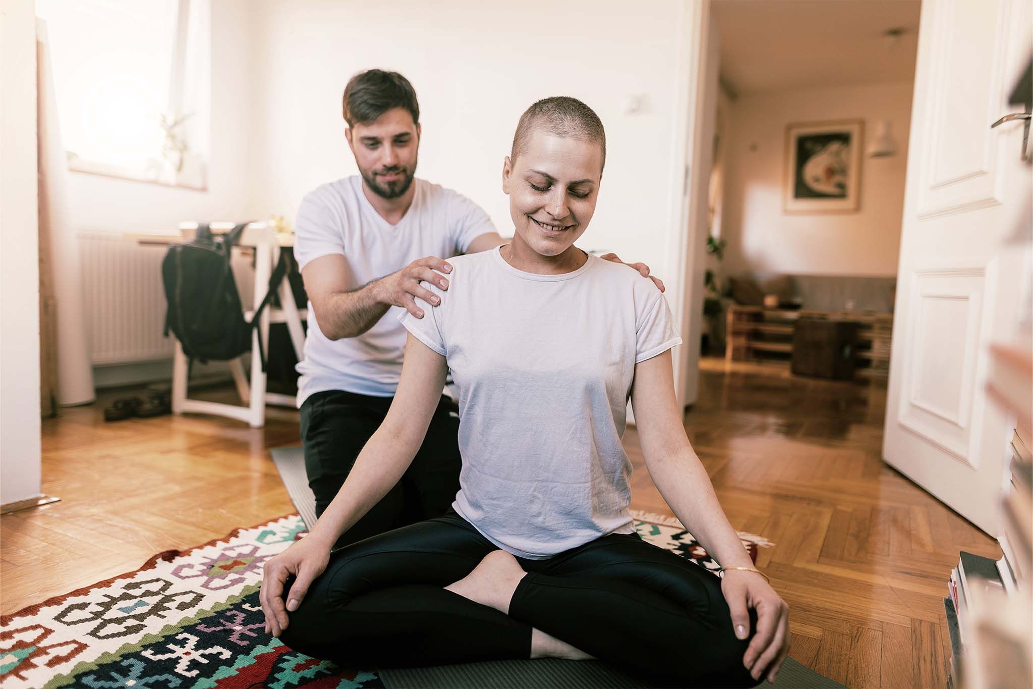 Eine Frau mit sehr kurzen Haaren sitzt lächelnd im Schneidersitz auf einem Teppich. Ihr Partner sitzt hinter ihr und hat ihr die Hände auf die Schultern gelegt.
