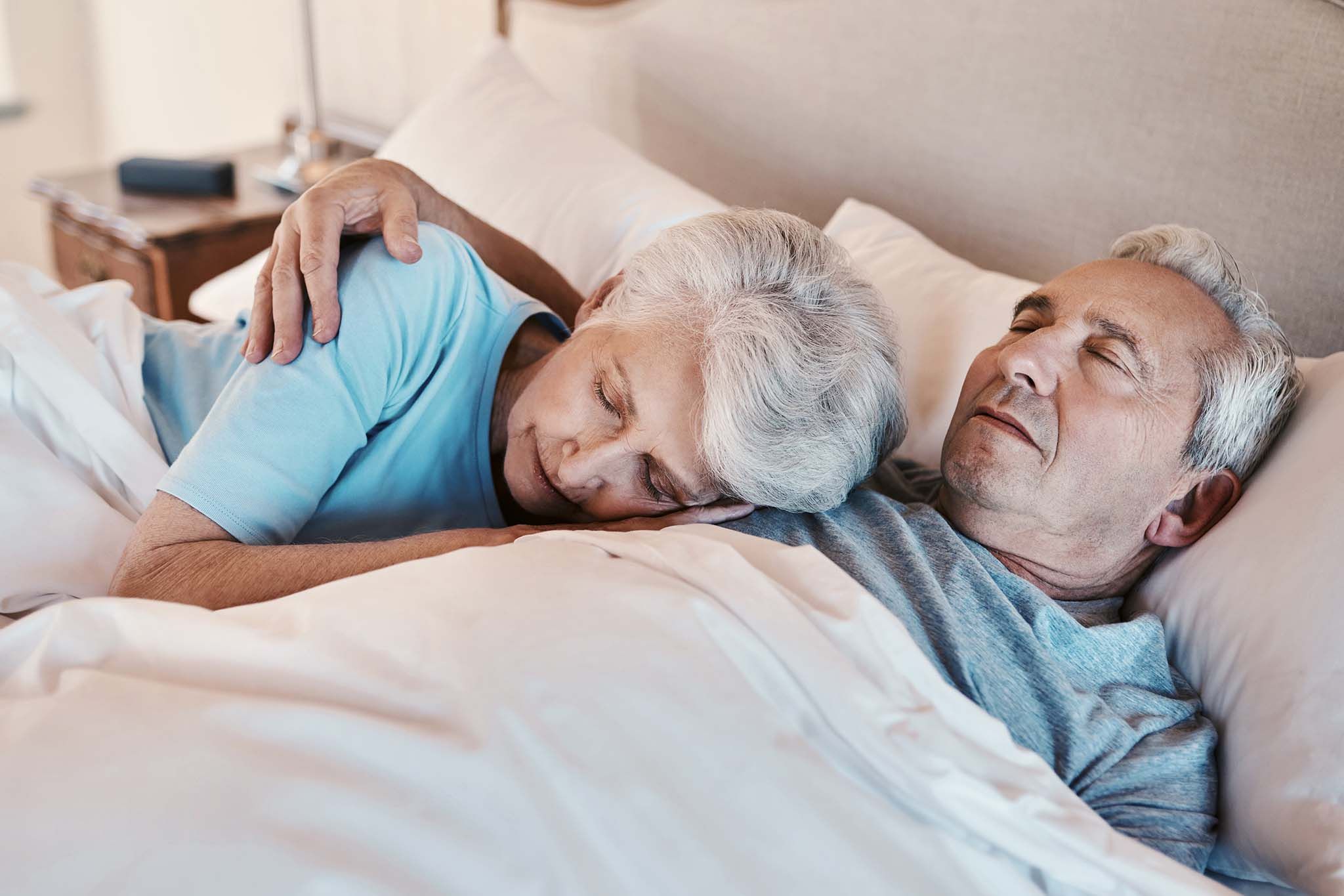 Ein älteres Paar liegt mit geschlossenen Augen im Bett. Die Frau legt ihren Kopf auf die Brust des Mannes, während er seinen Arm um ihre Schulter gelegt hat.
