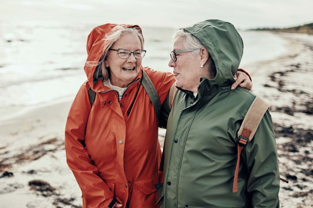 Zwei Frauen im Seniorenalter machen einen Spaziergang am Meer. Der Himmel ist wolkig, das Wetter kühl. Der Strand um sie herum ist menschenleer. Die beiden tragen Regenjacken, eine orange-rot, eine dunkelgrün, und Rucksäcke. Die Frau links in der rötlichen Jacke hat ihren Arm um ihre Freundin gelegt.