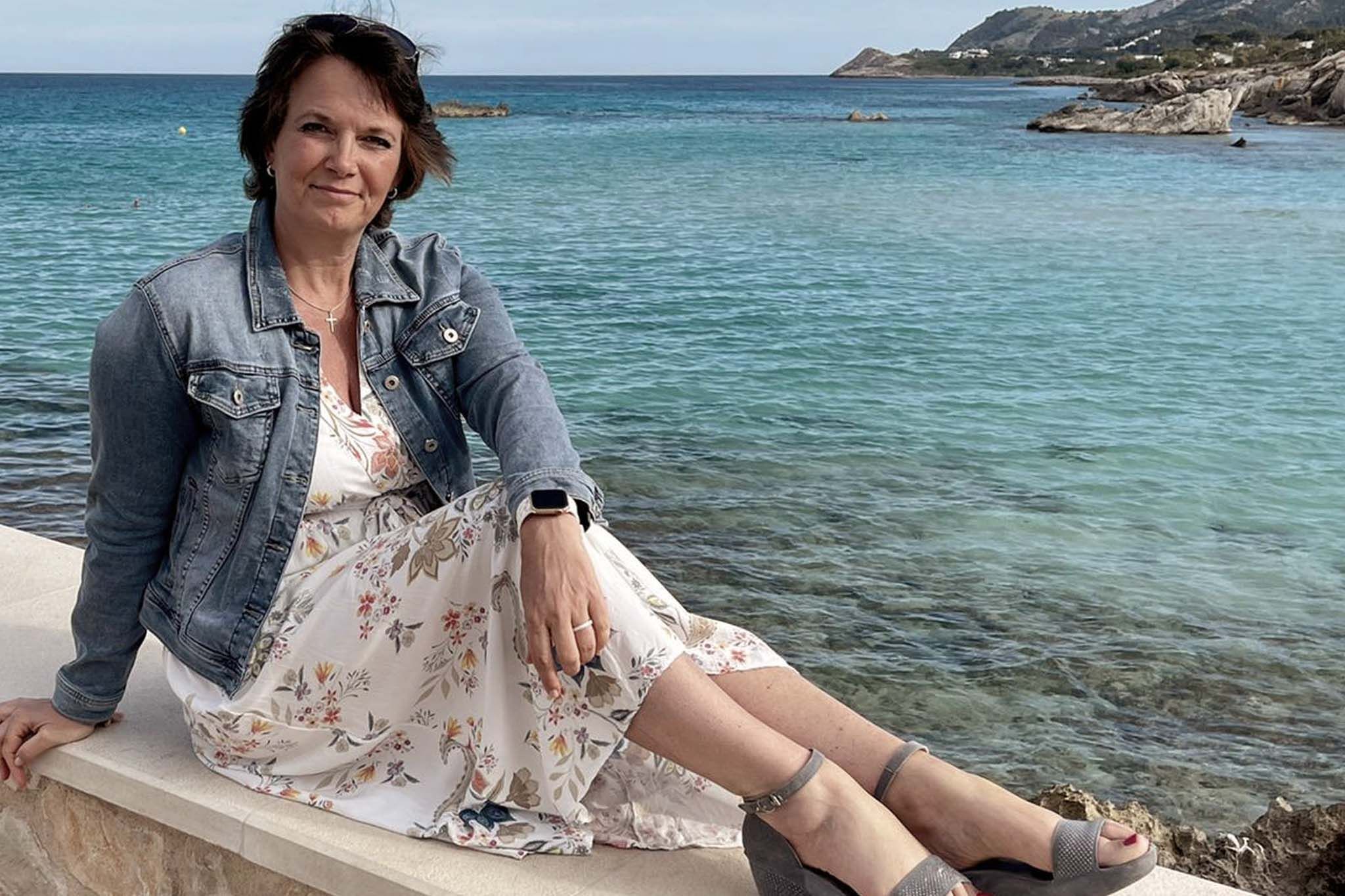 Martina sitzt entspannt auf einer Steinmauer am Meer und blickt in die Kamera. Sie trägt ein leichtes Sommerkleid und eine Jeansjacke. Im Hintergrund sind das klare Wasser und eine Küstenlandschaft zu sehen.