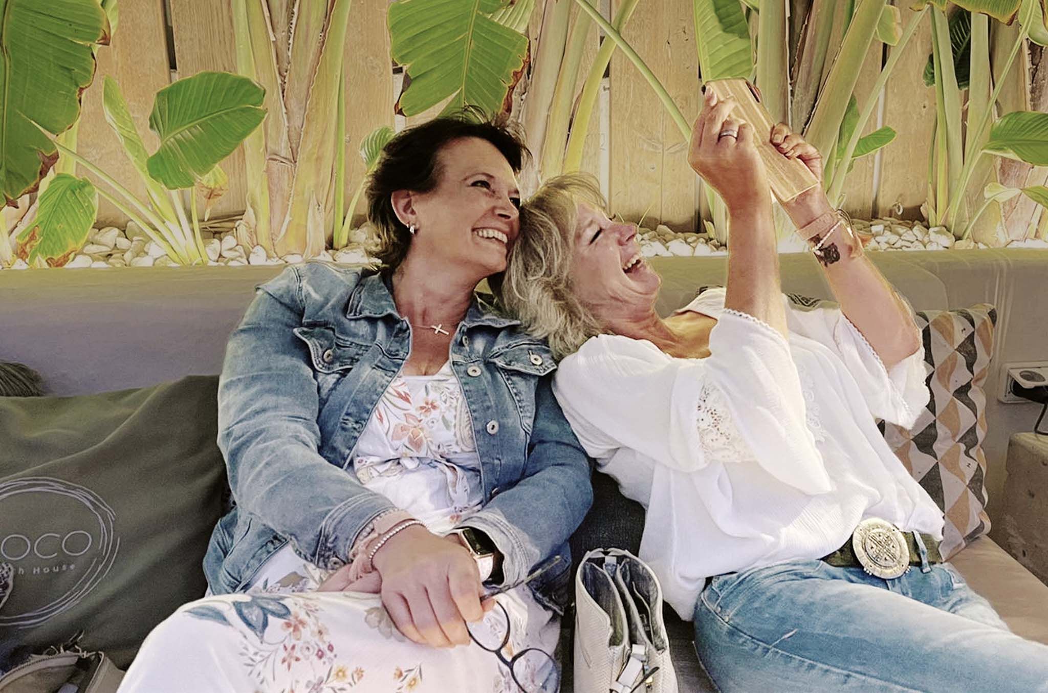 Martina sitzt mit einer anderen Frau entspannt auf einer Couch im Freien. Beide lachen und schauen gemeinsam auf ein Smartphone, während große grüne Pflanzen im Hintergrund zu sehen sind.