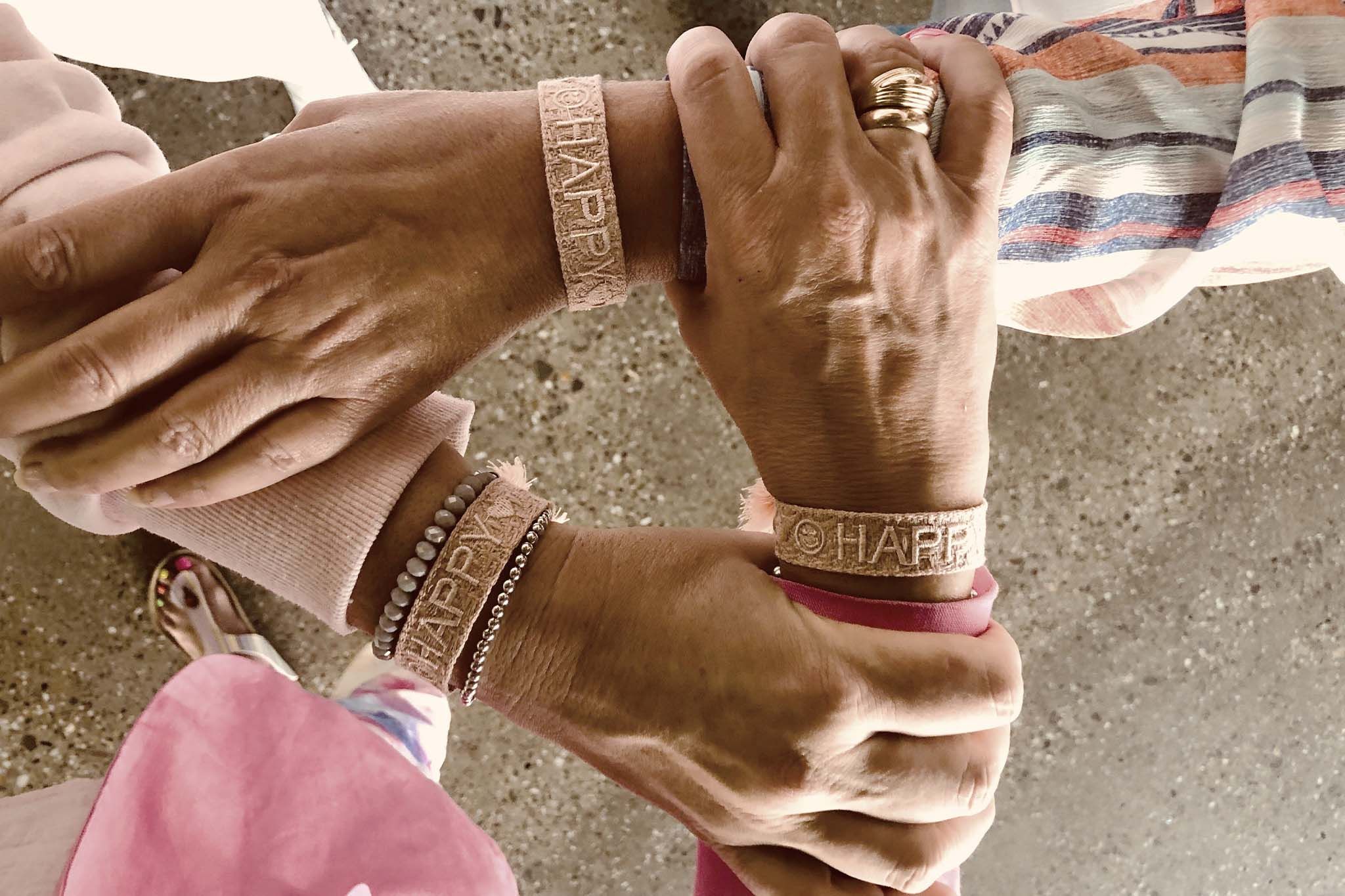 Martina legt gemeinsam mit drei weiteren Personen die Hände übereinander. Alle tragen Armbänder mit der Aufschrift „HAPPY“. Die Szene wirkt freundschaftlich und verbunden.