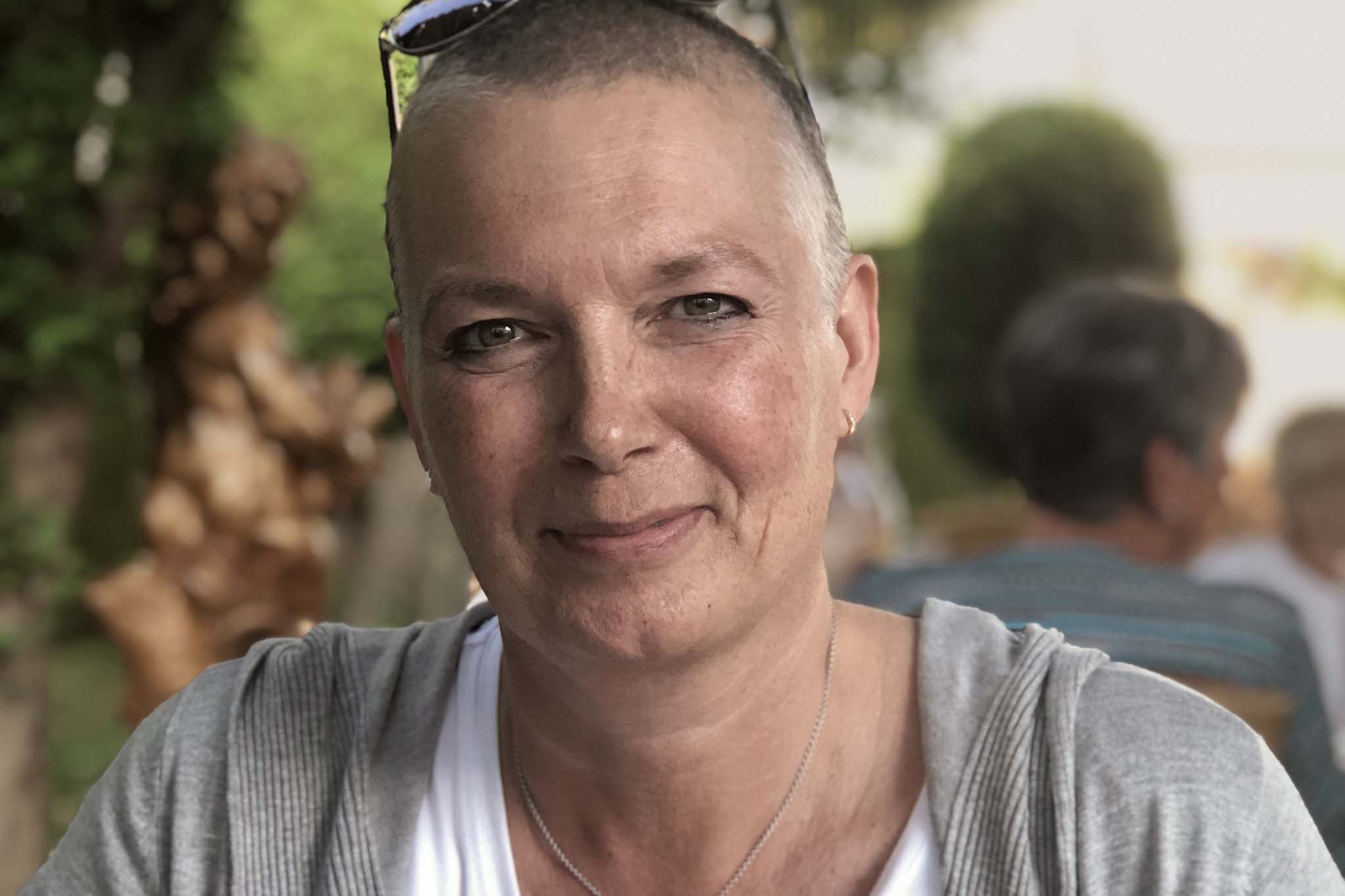 Martina sitzt im Freien an einem Tisch und blickt direkt in die Kamera. Sie trägt ein helles Oberteil, eine leichte Jacke und hat kurze Haare. Im Hintergrund sind unscharf Bäume und eine weitere Person zu sehen.