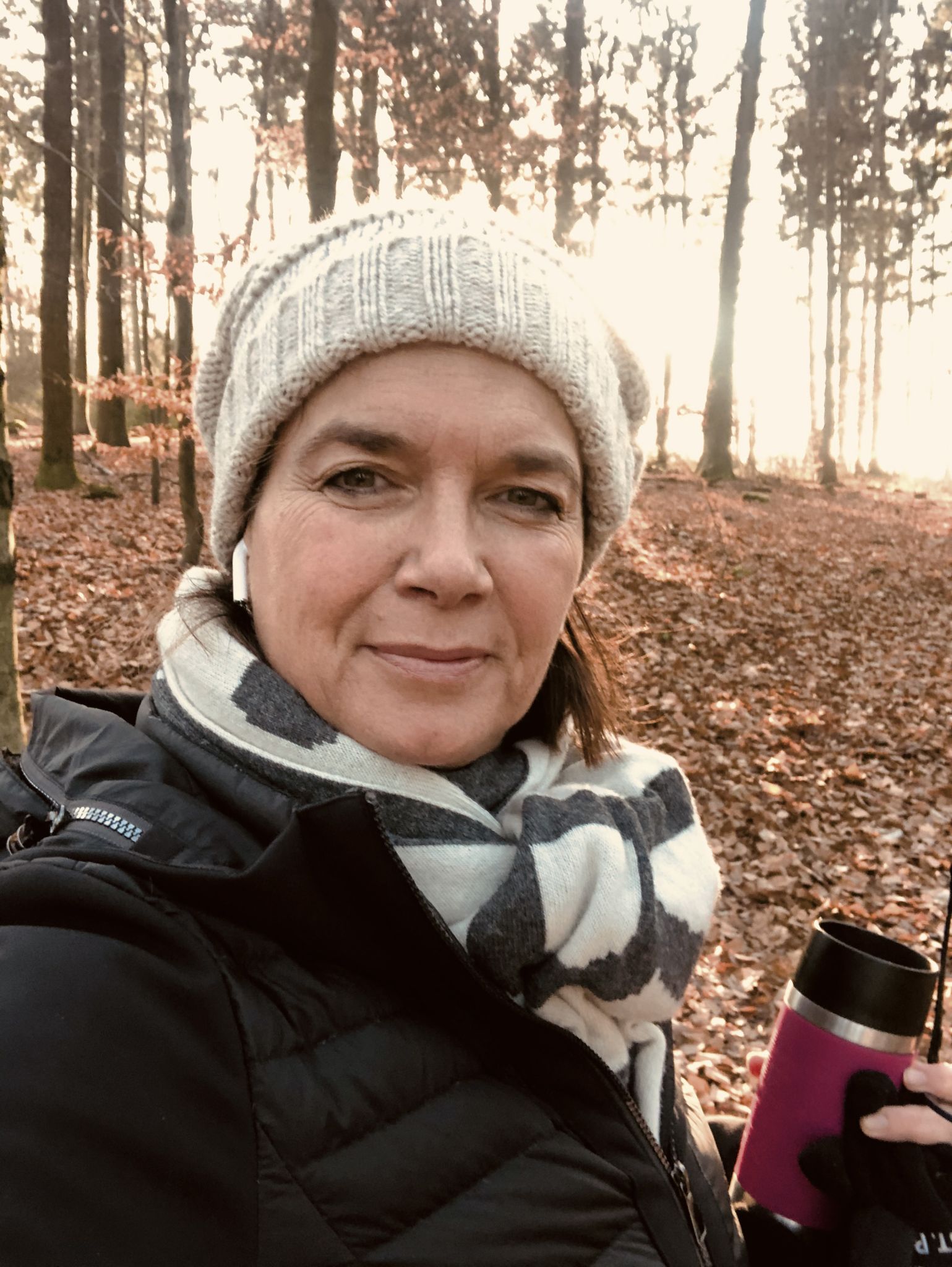 Martina steht in einem herbstlichen Wald und blickt in die Kamera. Sie trägt eine Strickmütze, einen Schal und eine Jacke. Der Boden ist mit Laub bedeckt, und durch die Bäume fällt warmes Sonnenlicht.