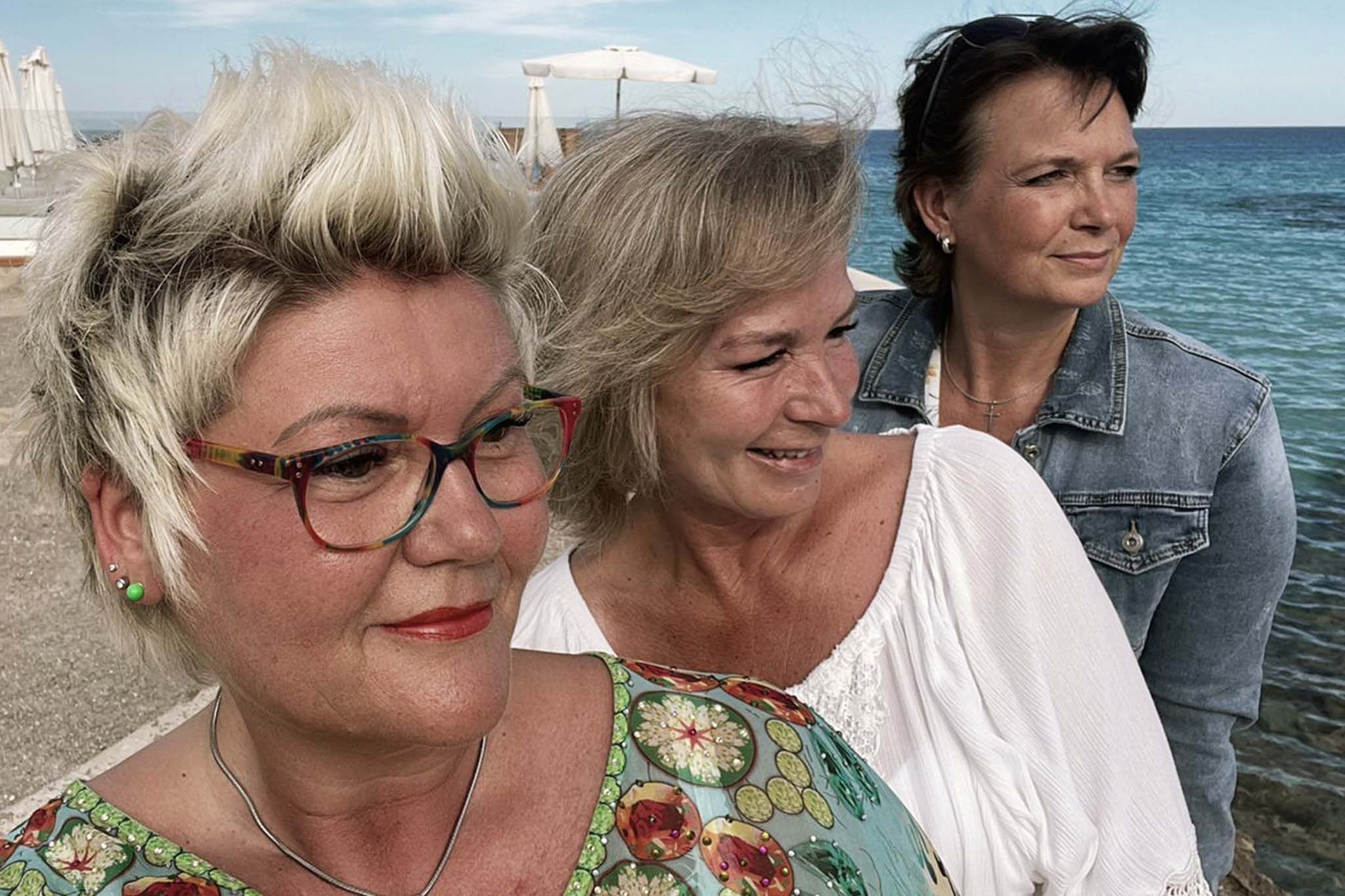 Martina steht mit zwei weiteren Frauen am Strand und blickt gemeinsam mit ihnen aufs Meer. Im Hintergrund sind Wasser, Strand und Sonnenschirme zu sehen. Die Stimmung wirkt entspannt und sommerlich.