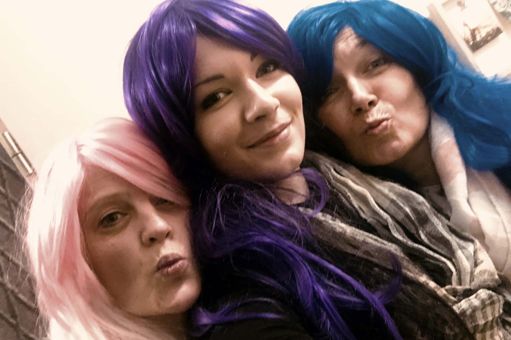 Martina und zwei Freundinnen machen ein gemeinsames Selfie. Sie tragen bunte Langhaar-Perücken: eine rosafarbene, eine violette und eine blaue. Amüsiert blicken sie in die Kamera.
