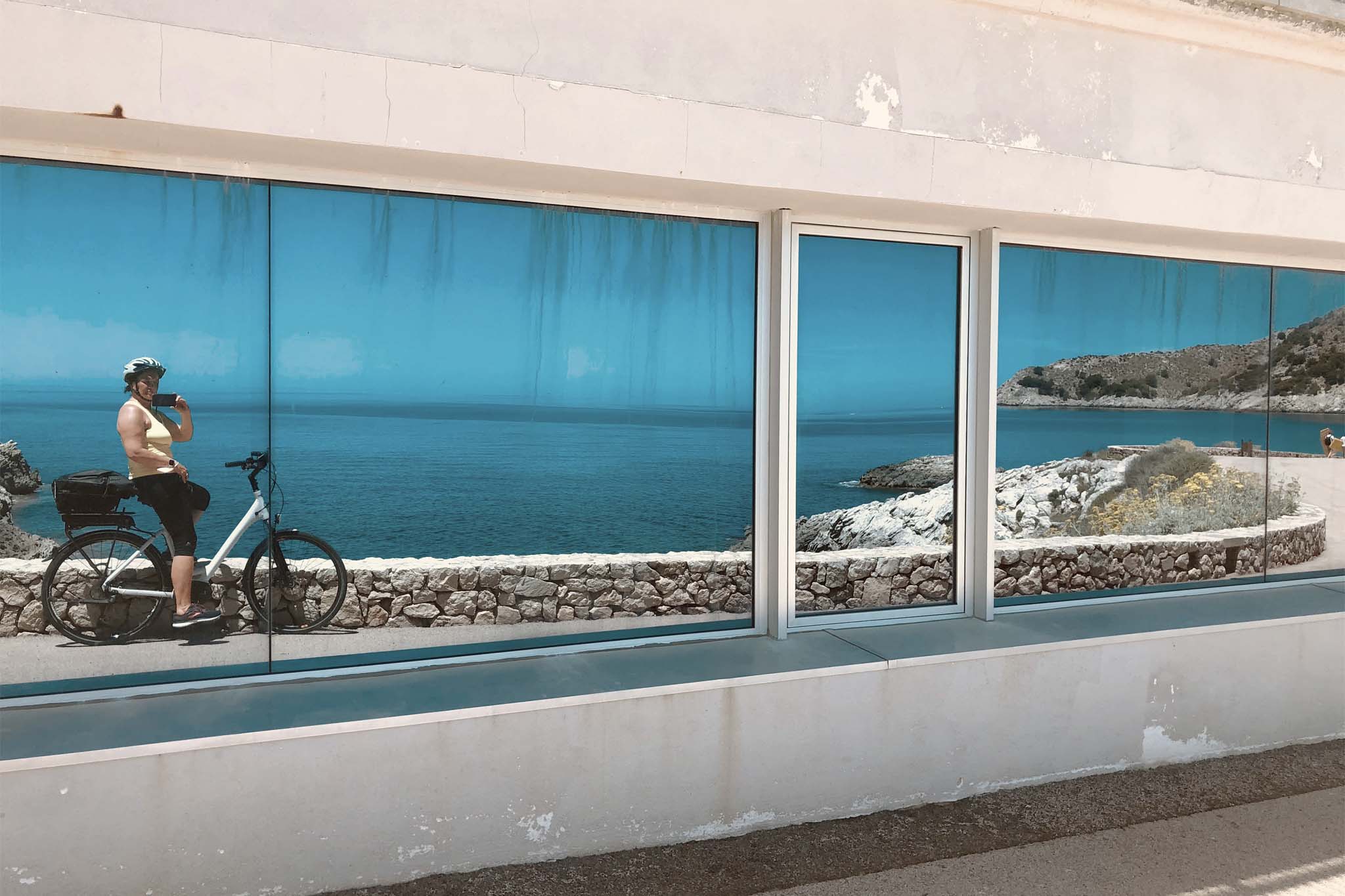 An einer weißen Häuserfassade ist ein Spiegel angebracht. In der Spiegelung ist eine Radfahrerin, Martina, zu sehen, die von sich ein Foto im Spiegel macht, die inmitten traumhafter Natur eine Pause macht. Hinter dem Radwanderweg liegen Meer und Felsen.