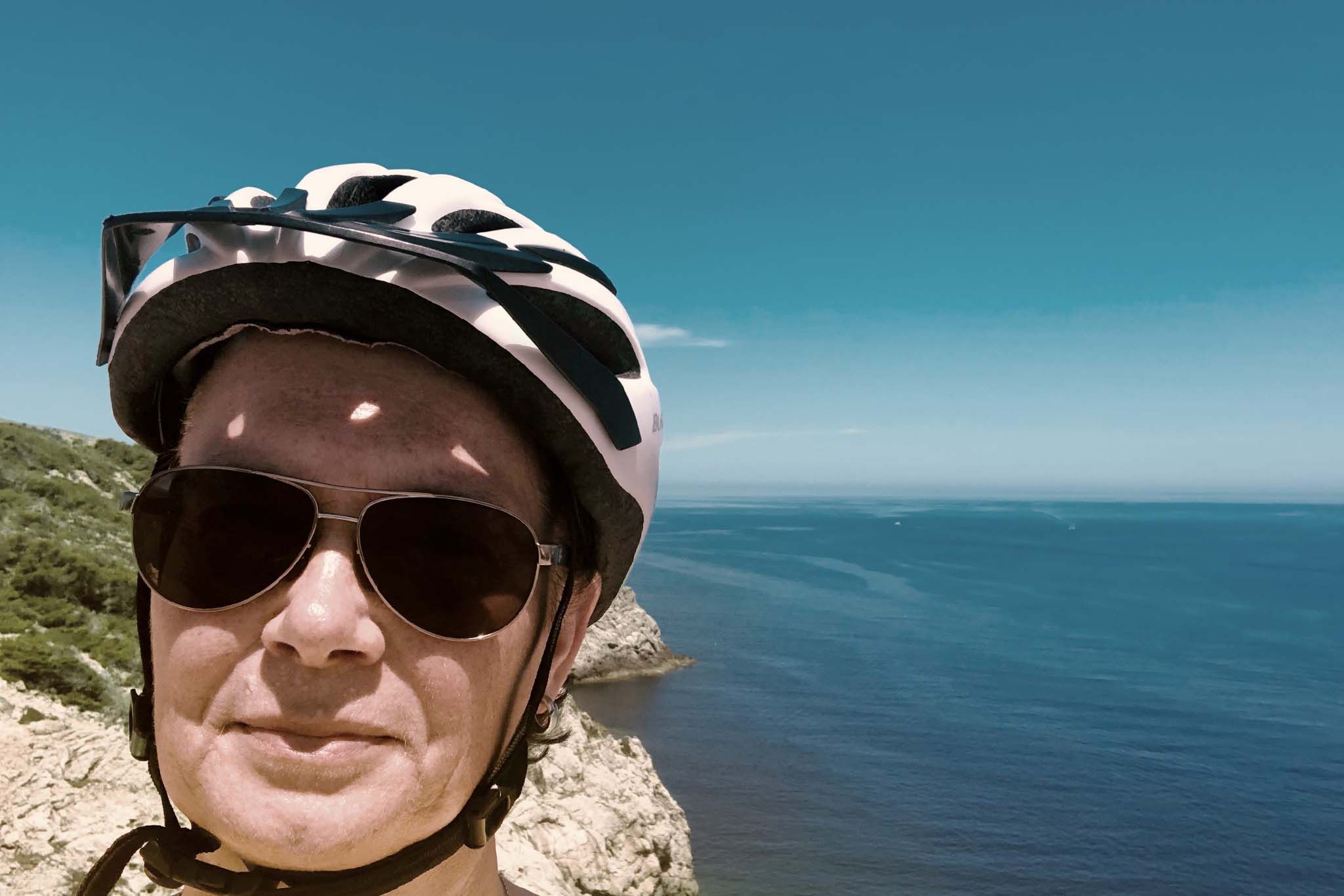 Martina steht mit Fahrradhelm und Sonnenbrille an einer Küste und blickt in die Kamera. Hinter ihr sind das Meer, Felsen und ein klarer blauer Himmel zu sehen.