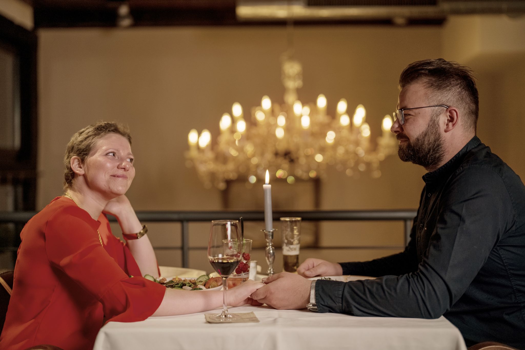 Silja und ihr Mann sitzen an einem Tisch im Restaurant, blicken sich an und halten sich an den Händen. Im Hintergrund ist ein Kronleuchter zu sehen.