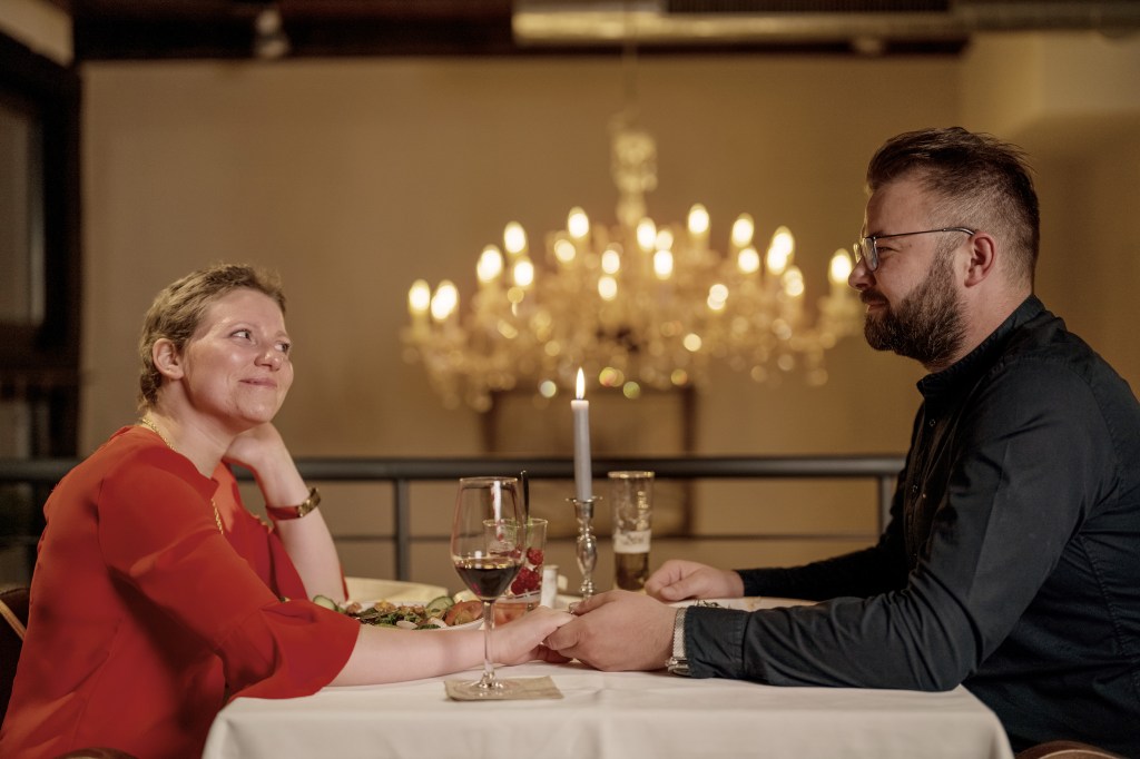 Silja und ihr Mann sitzen in einem Restaurant. Sie trägt ein rotes Kleid und er ein schwarzes Hemd. Ein edler Kronleuchter leuchtet hinter ihnen im romantischen Halbdunkel. Die beiden blicken sich verliebt an. Ihre Hände berühren sich auf dem Tisch.