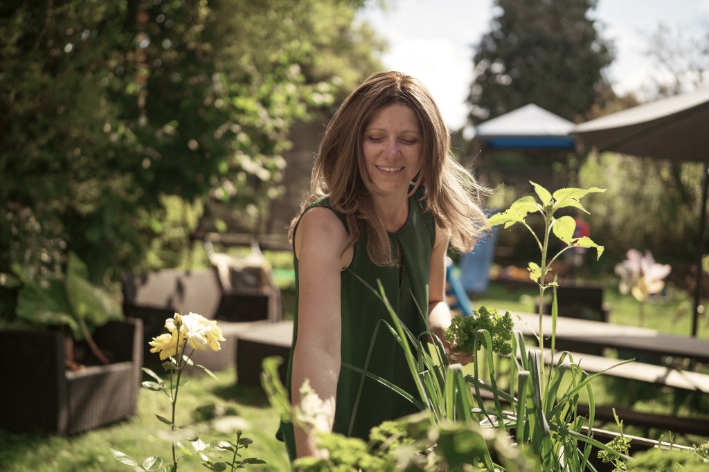 Silja steht vor einem Hochbeet in ihrem Garten. Lächelnd pflückt sie etwas und genießt das Sommerwetter. Sie trägt die Haare lang und offen.