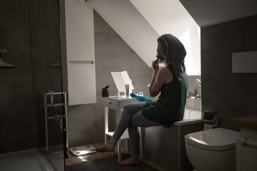 Silja sitzt auf dem Rand ihrer Badewanne im Halbdunkel und trägt sich eine Gesichtsmaske auf. Durch das Dachfenster fällt etwas Licht auf ihr Profil.