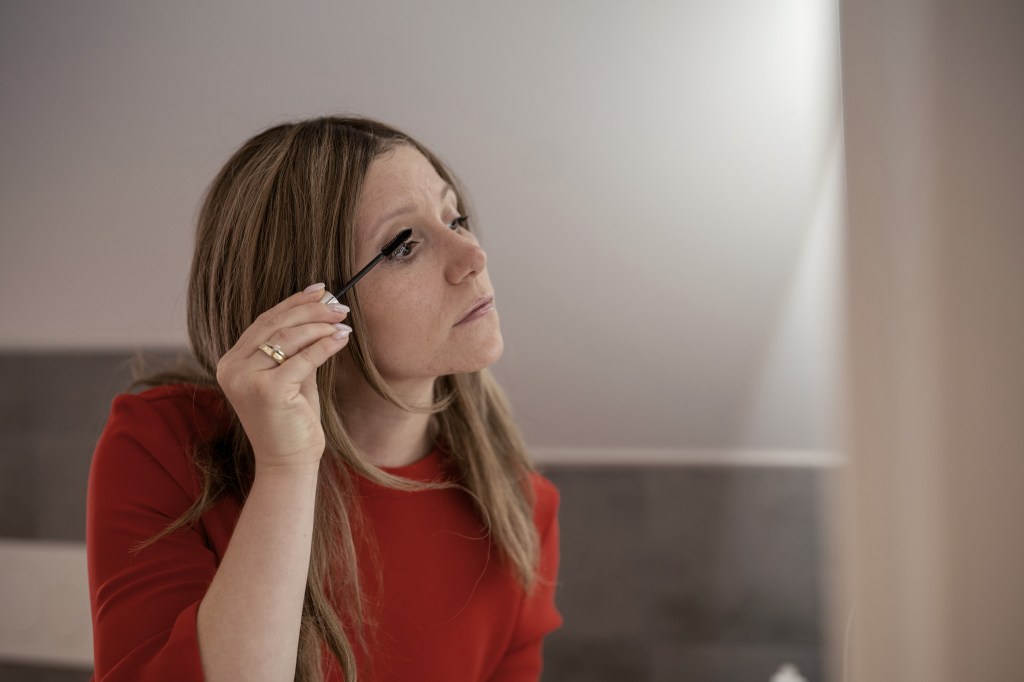 Silja trägt mit konzentriertem Blick Mascara auf. Sie hat ein rotes Kleid an und trägt die Haare offen und lang.