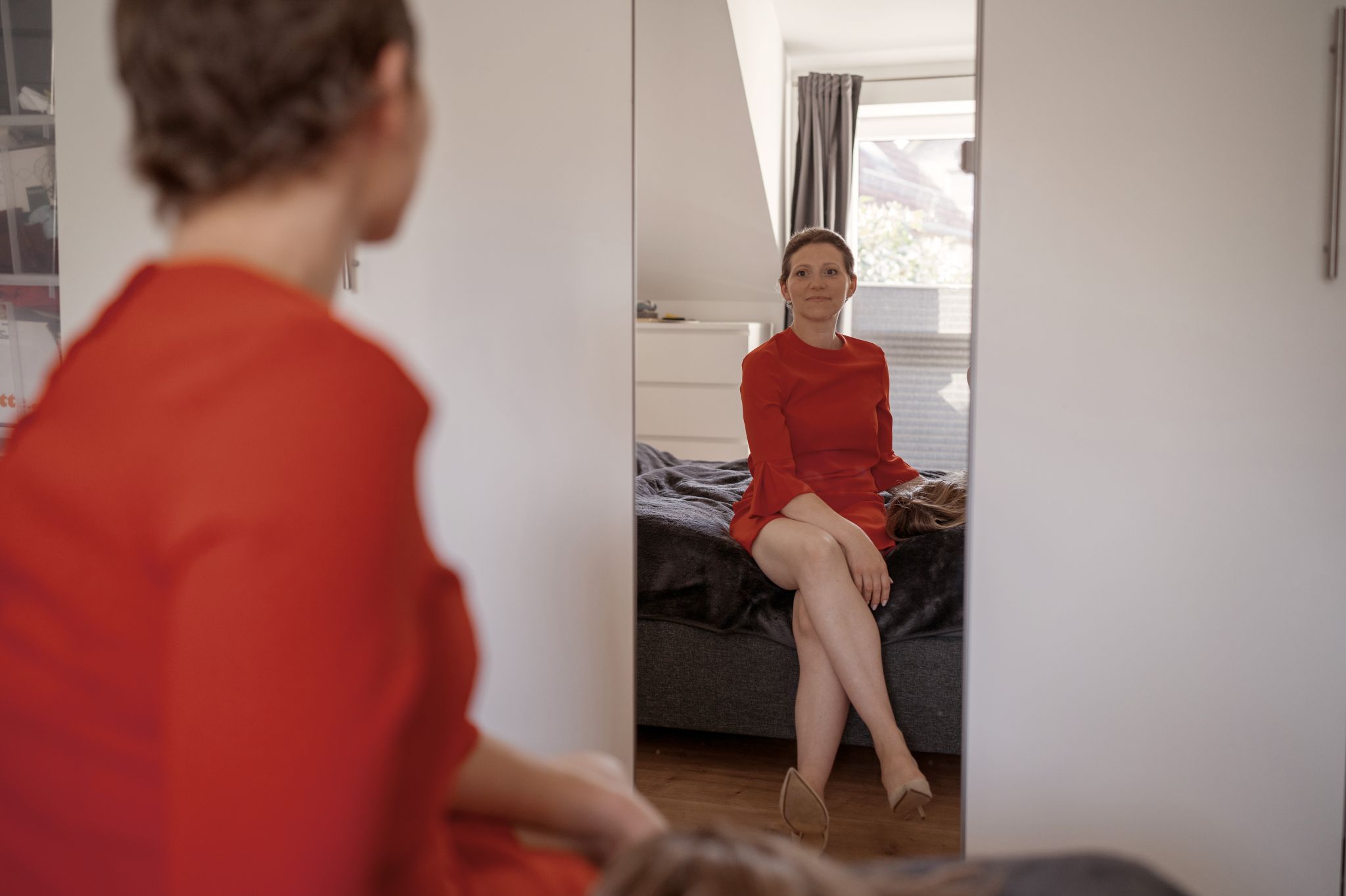 Silja sitzt in einem roten Kleid auf ihrem Bett vor dem Spiegel ihres Kleiderschranks und sieht sich zufrieden an. Sie hat ihre Perücke neben sich gelegt und man kann ihre braune Kurzhaarfrisur sehen.
