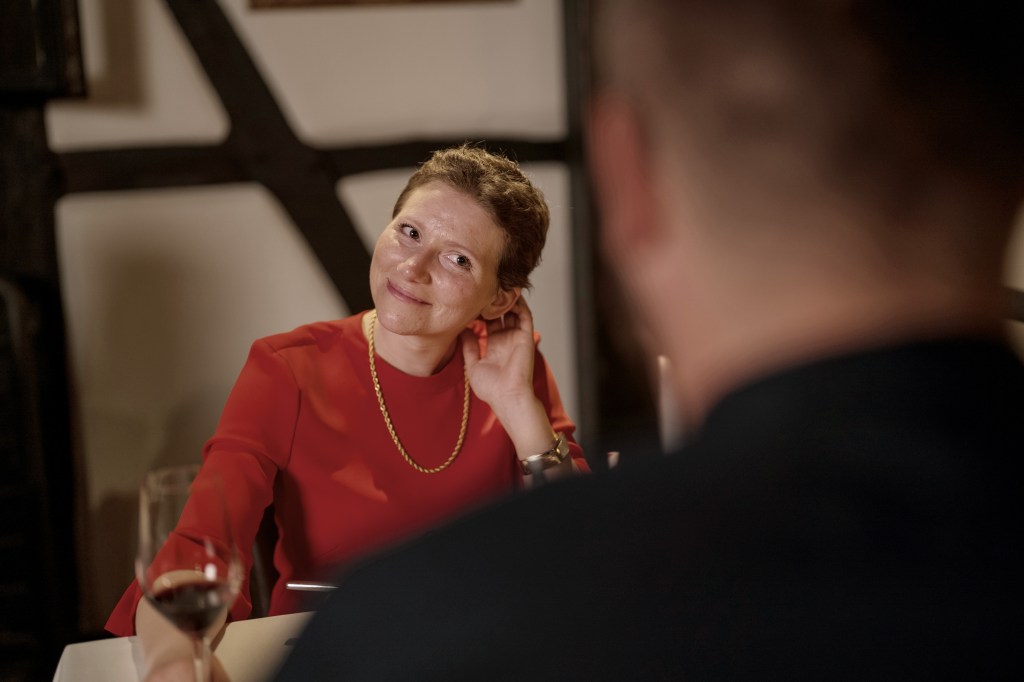 Silja besucht mit ihrem Mann ein Restaurant. Sie sitzen einander gegenüber und trinken Wein. Silja hat ein rotes Kleid an und trägt die Haare ganz kurz. Ein wenig nervös lächelt sie ihren Mann an.