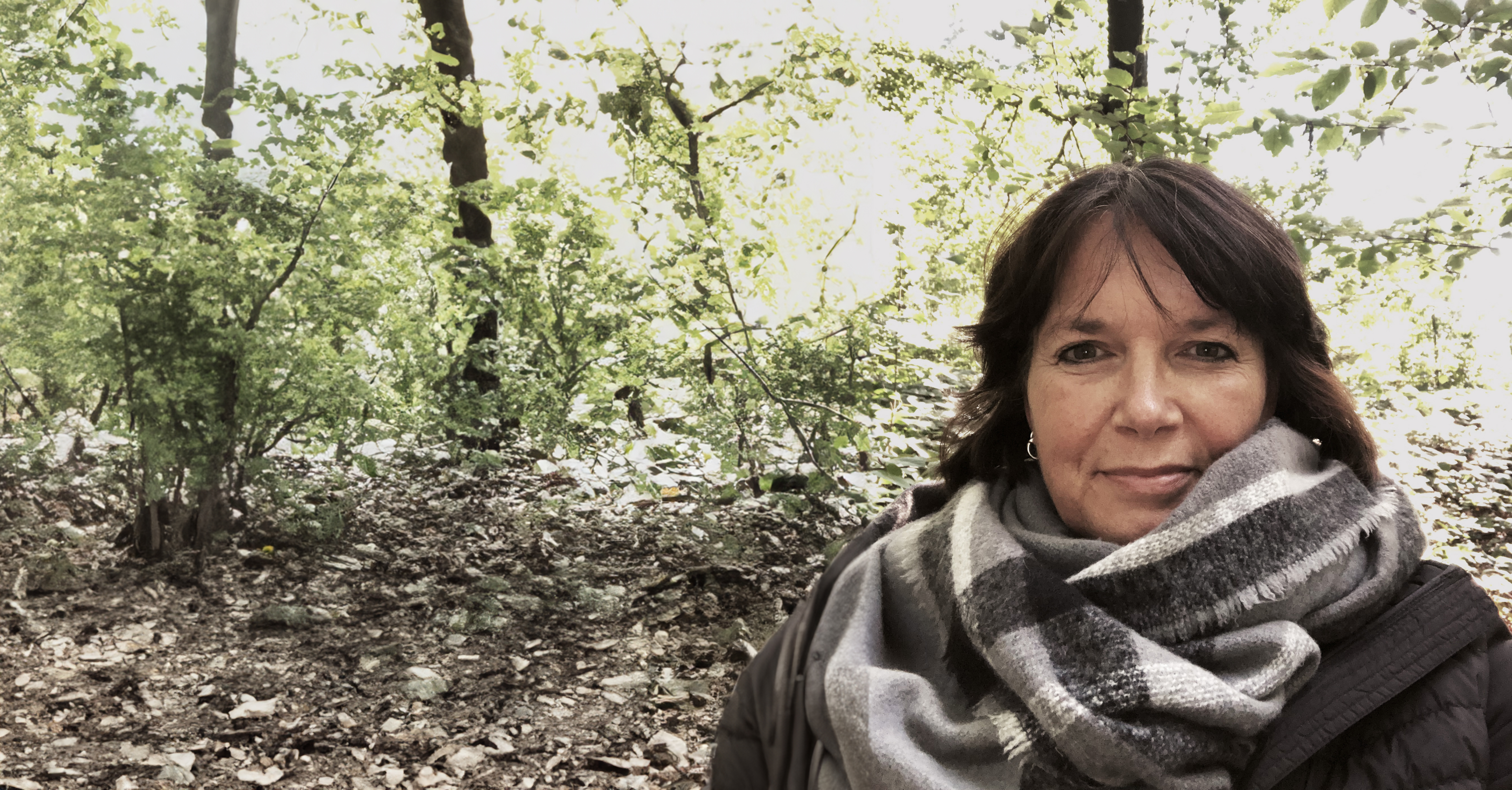 Martina blickt lächelnd in die Kamera. Sie trägt einen grauen Schal und ist umgeben von Wald.