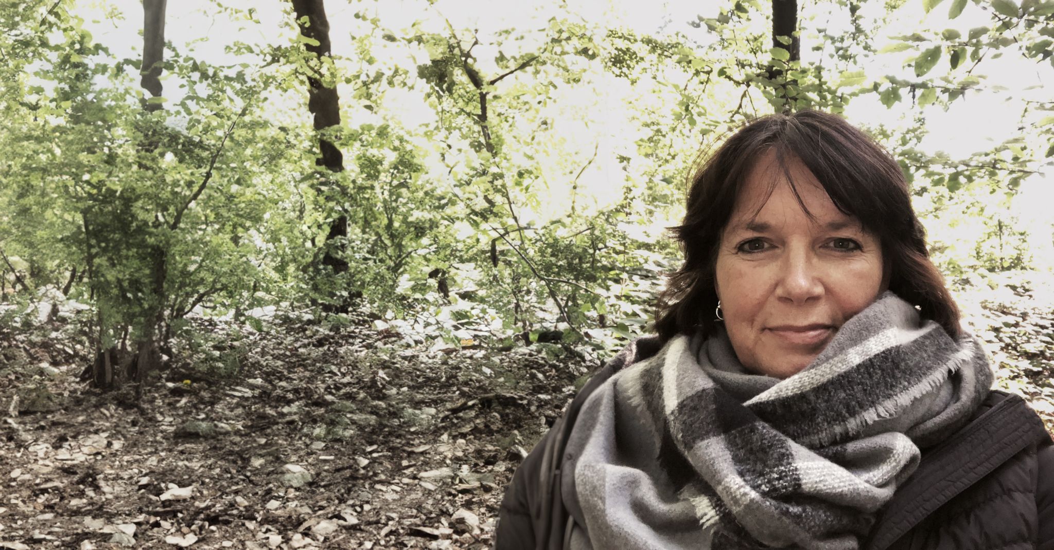 Martina steht im Wald und blickt ruhig in die Kamera. Sie trägt einen dicken Schal und eine Jacke. Um sie herum sind Bäume und herbstliches Laub zu sehen.
