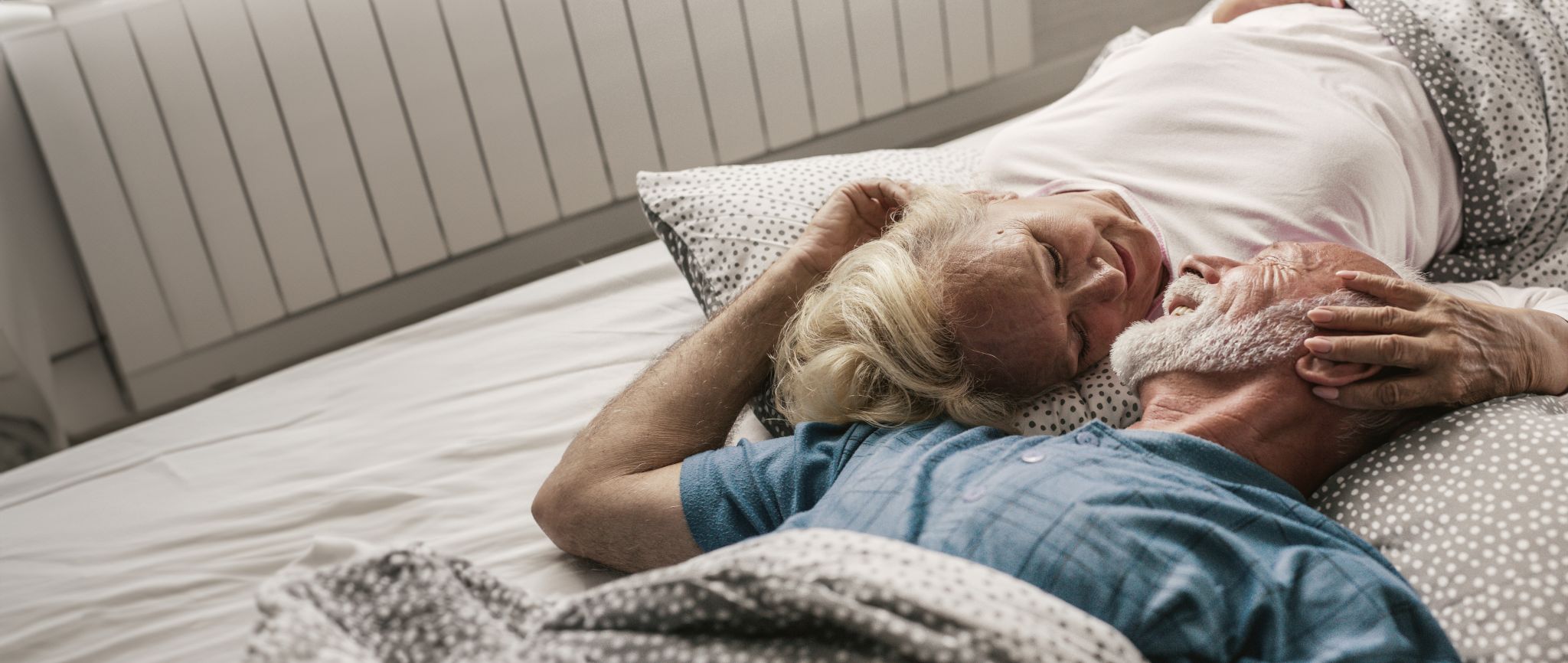 Ein älterer Mann und eine ältere Frau liegen auf einem Bett und haben ihre Gesichter zueinander gewandt.