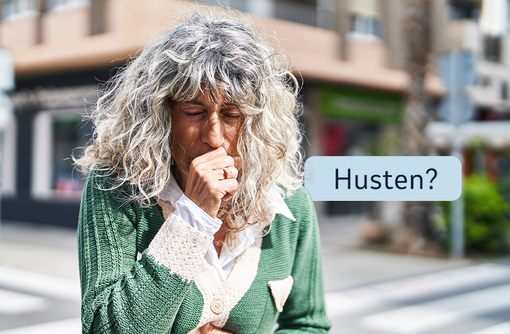 Eine Frau mit grauen, lockigen Haaren steht im Freien in einer städtischen Umgebung. Sie hält ihre Hand vor den Mund, während sie hustet. Über ihr erscheint eine Sprechblase mit dem Wort „Husten?“. Im Hintergrund sind unscharfe Gebäude und eine Straße zu sehen.